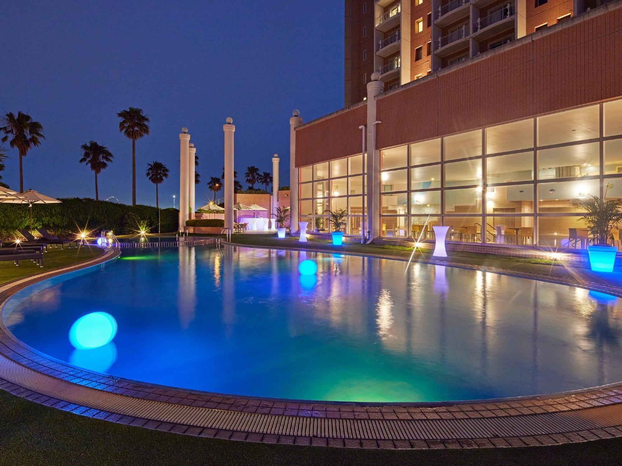 מלון Urayasu Grand Nikko Tokyo Bay Maihama מראה חיצוני תמונה The photo shows a well-lit swimming pool at night, surrounded by a lush green area. The pool features glowing blue lights that create a serene and inviting atmosphere. Palm trees can be seen in the background, adding a tropical feel to the scene. Add